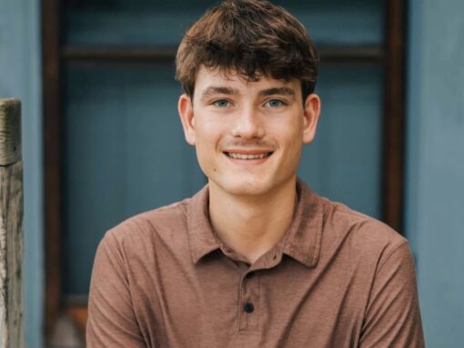 A young man with short brown hair and blue eyes, wearing a brown collared shirt, sits in front of a blue wall and looks at the camera with a slight smile.