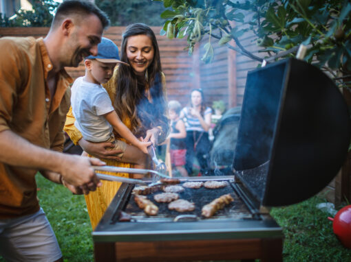 A group of people barbecue meat outdoors in a backyard, enjoying the day with mosquito control for homeowners ensuring a comfortable gathering. A man grills, a woman holds a child, and others mingle in the background.