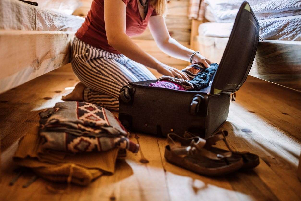 Person packing clothes into a suitcase on a wooden floor beside a bed
